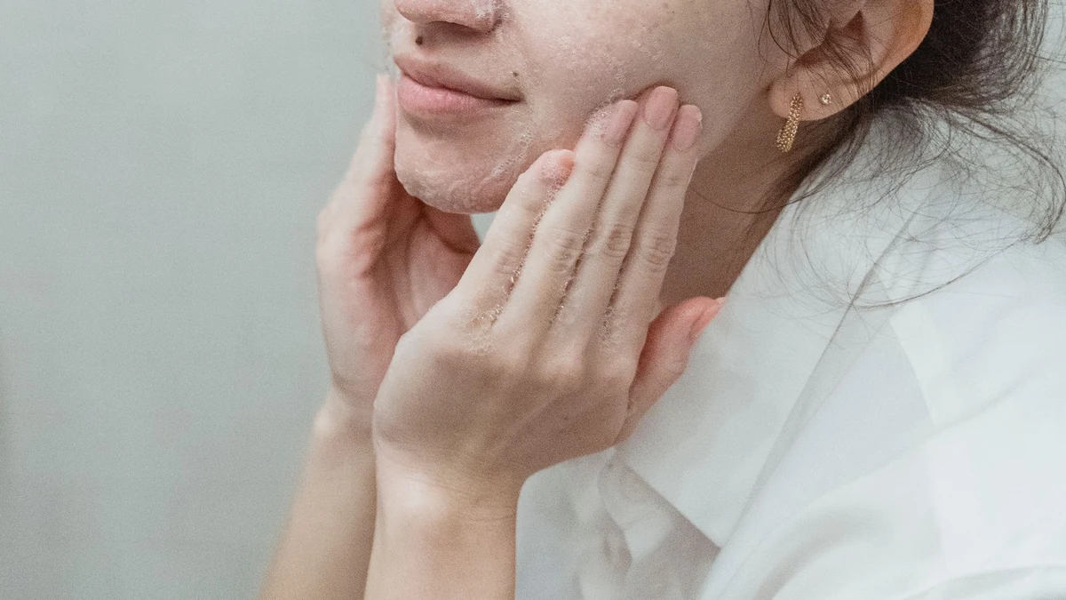 Woman gently massaging foaming exfoliating cleanser onto cheeks at bathroom washbasin